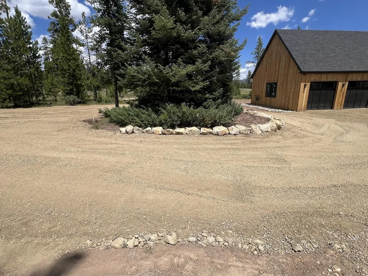 A boulder-ringed spruce in the middle of a graded gravel drive beside a cedar garage