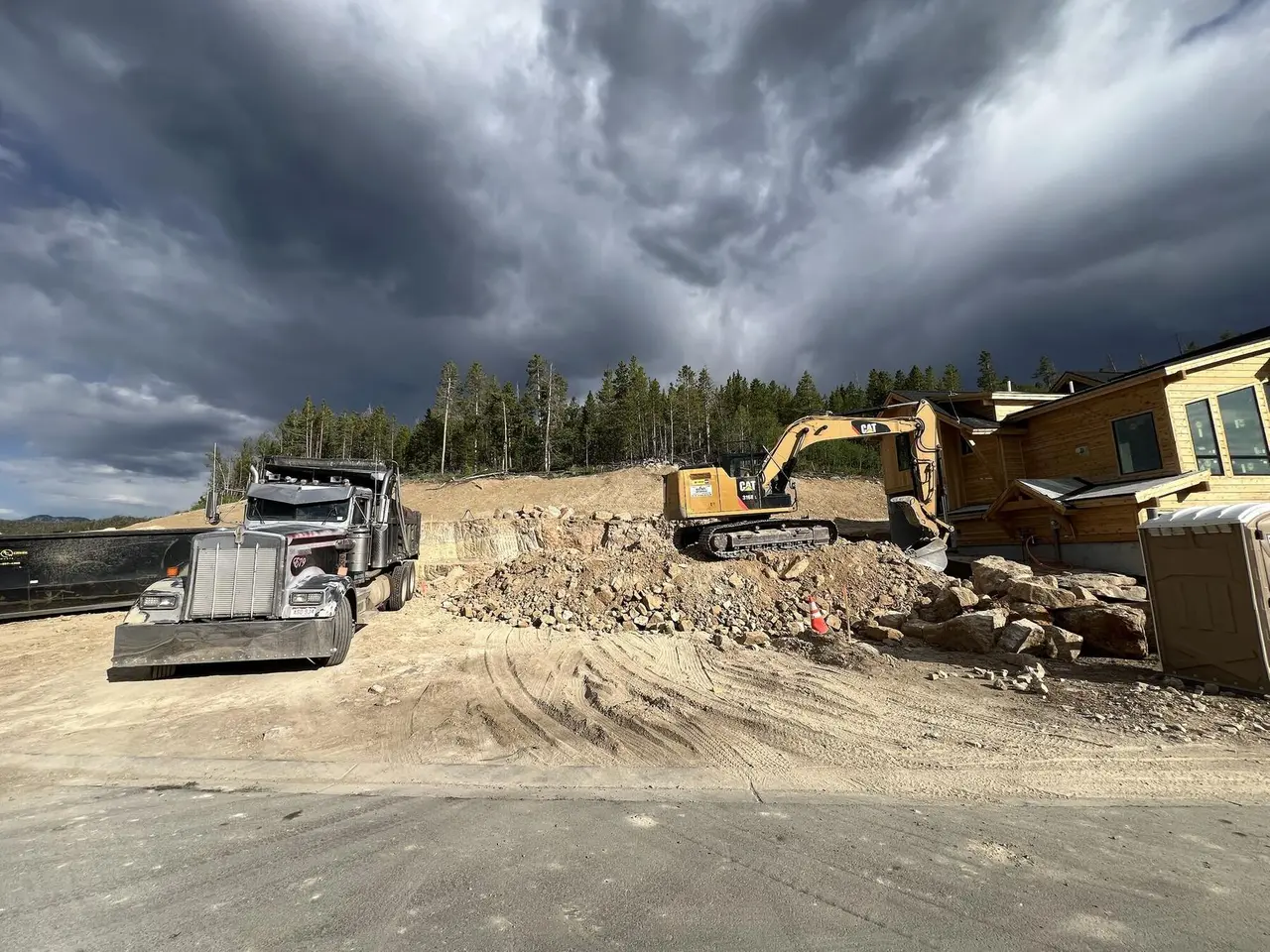 A dump truck and an excavator beside a pile of broken rock under storm clouds