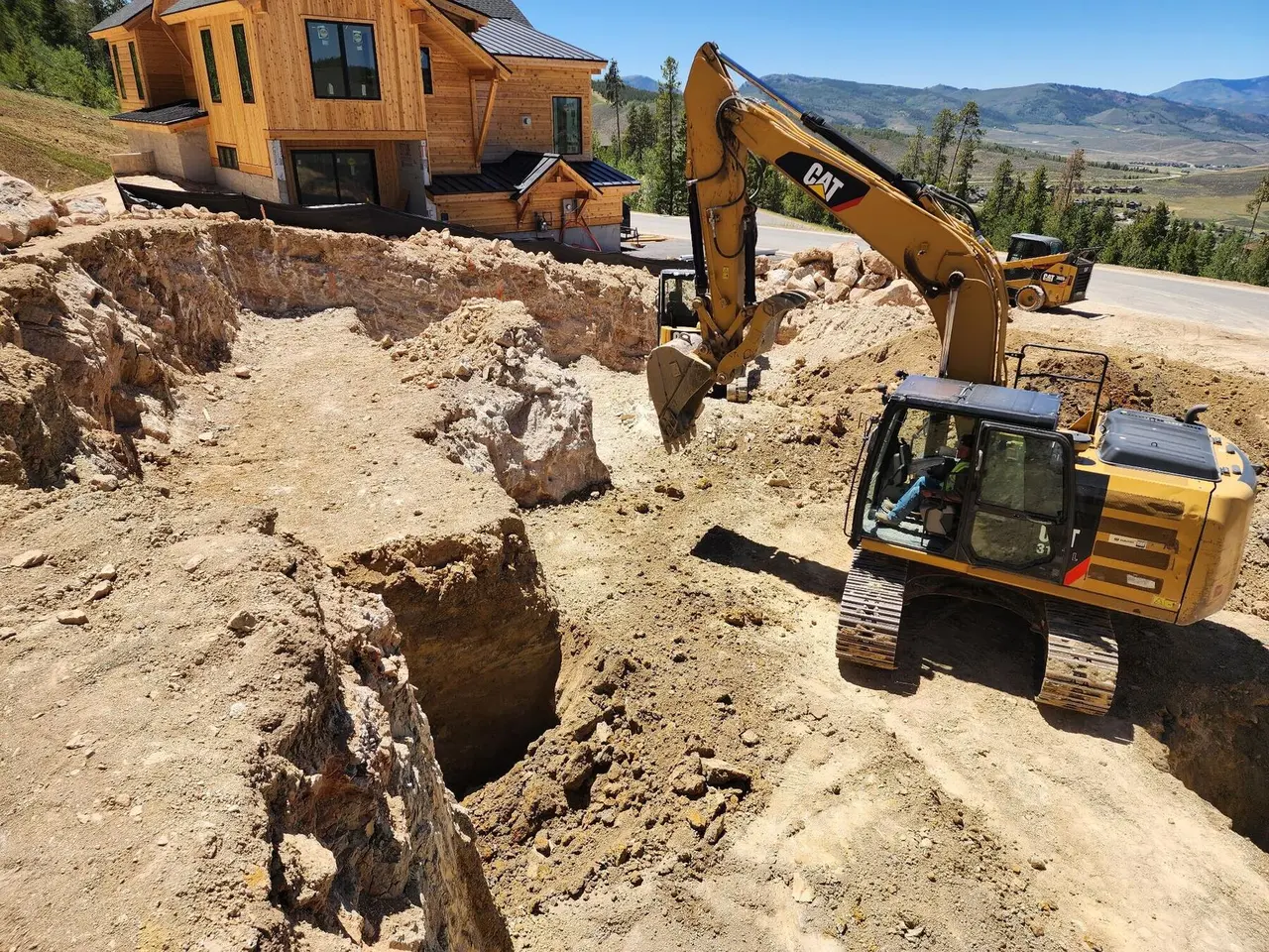 An excavator digging a home foundation on a hillside above a mountain valley