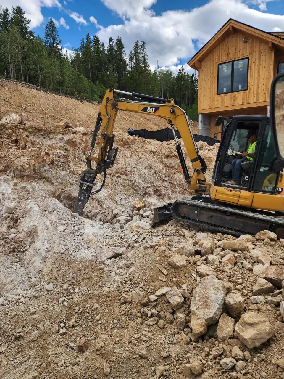 A mini excavator with a hydraulic hammer breaking bedrock beside a new cedar home