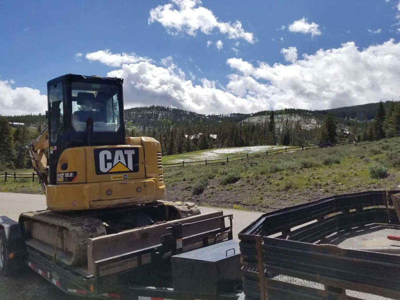 A CAT mini excavator strapped to a trailer below a forested hill