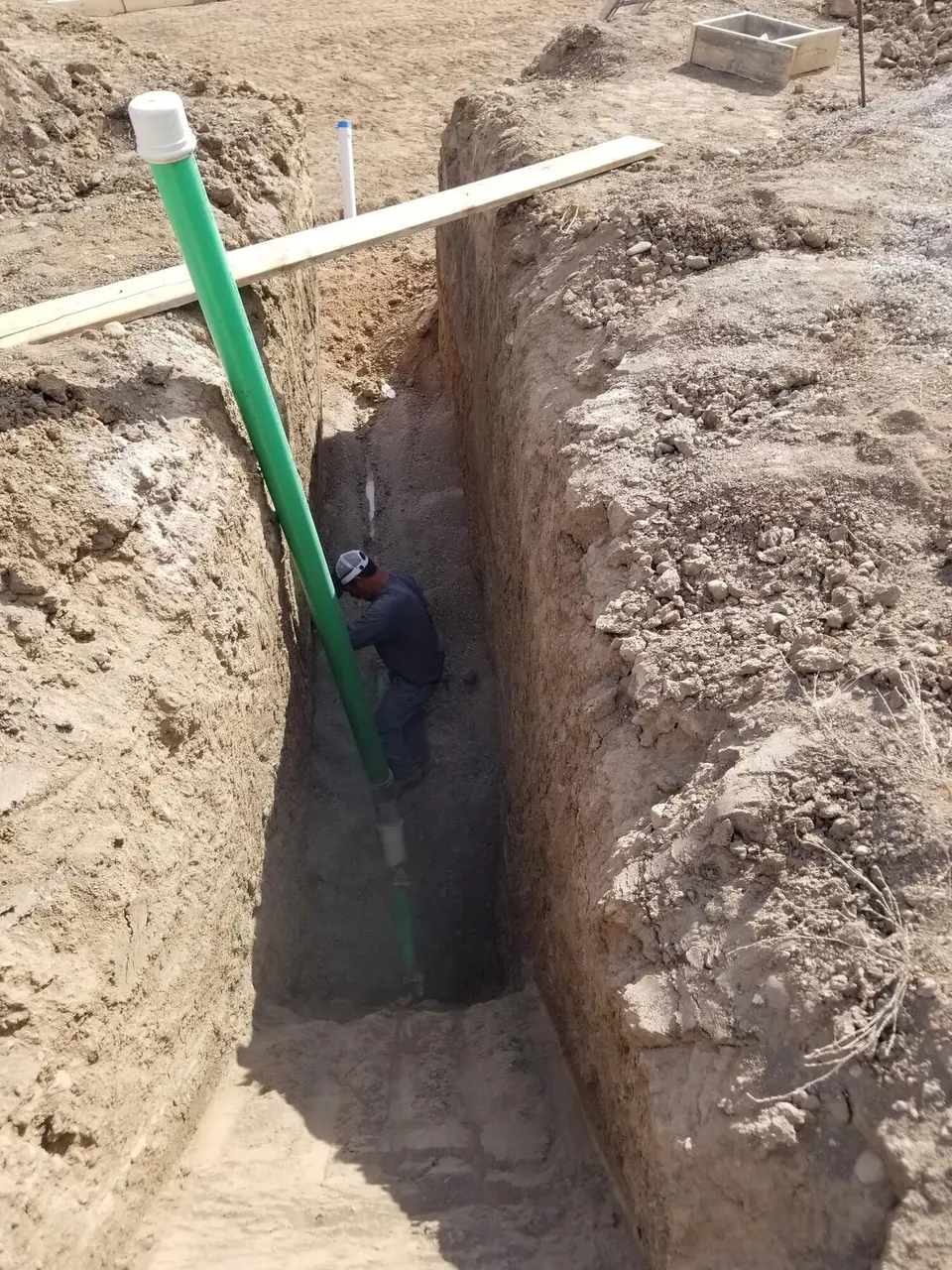 A crew member working at the bottom of a deep trench beside a green sewer pipe