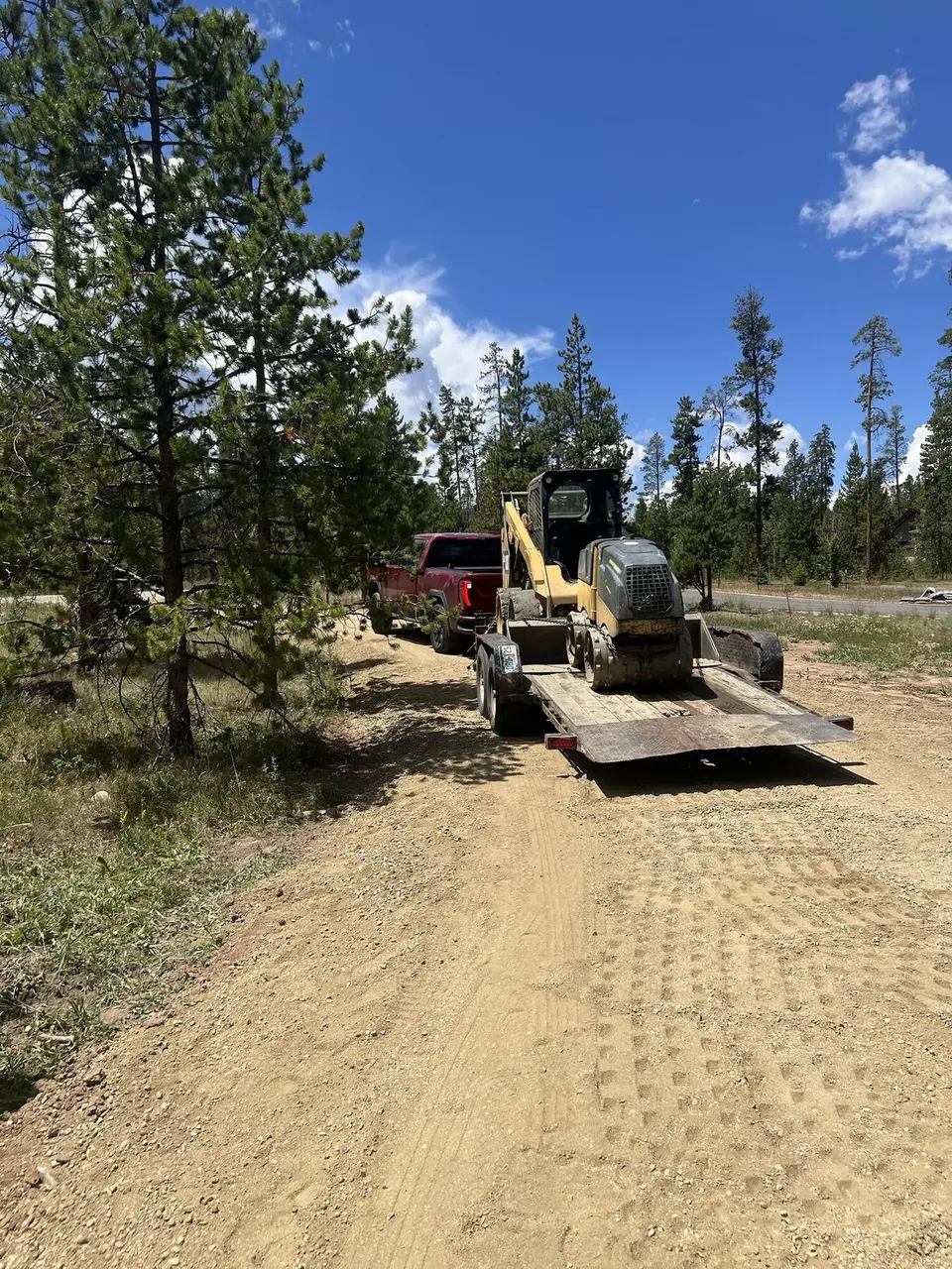 A tracked skid steer on a trailer behind a red pickup on a forest road